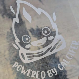 May include: A white vinyl decal featuring a stylised flame character with large circular eyes and a small mouth, positioned above a log. The text "POWERED BY CALCIFER" is printed in a curved, sans-serif font beneath the flame illustration.