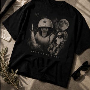 May include: A black t-shirt featuring a graphic print of a chimpanzee wearing a NASA helmet, a rocket launching, a full moon, and a chimpanzee sitting inside a space capsule. The text "SPACE CHIMP LIVES" is printed in white at the bottom of the graphic.