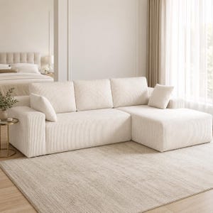 May include: A cream-colored sectional sofa upholstered in a textured corduroy fabric. The sofa features three back cushions, a chaise lounge section on the right side, and two matching square throw pillows positioned on the left and right ends.