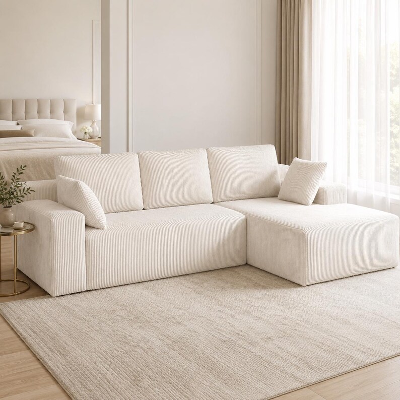 May include: A cream-colored sectional sofa upholstered in a textured corduroy fabric. The sofa features three back cushions, a chaise lounge section on the right side, and two matching square throw pillows positioned on the left and right ends.
