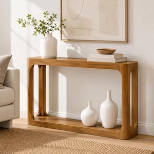 May include: A rectangular wooden console table with rounded inner corners, featuring a lower shelf. On the top surface sits a white ceramic vase with green foliage and a stack of two books topped with a small wooden bowl. Two white vases sit below.