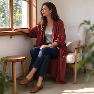 Könnte beinhalten: Eine lange, offen zu tragende Strickjacke in Rostrot mit langen Ärmeln, kombiniert mit einem hellgrauen Tanktop und einer dunkelblauen Jeans. Das Outfit wird durch ein Paar braune Leder-Loafer ergänzt, was einen Lagen-Look in neutralen Tönen ergibt.