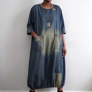 May include: A midi-length denim dress featuring a round neckline, three-quarter sleeves, and two large front pockets. The fabric has a faded blue wash with distressed olive-toned patches on the shoulders, cuffs, and hem, finished with a gathered elastic bottom.