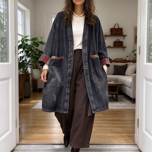 Puede incluir: Un abrigo largo de mezclilla gris carbón con cierre de botones, dos bolsillos delanteros grandes y puños doblados con un patrón de cuadros en rojo, negro y beige. El abrigo se usa sobre una blusa color crema de manga larga y pantalones marrón oscuro.