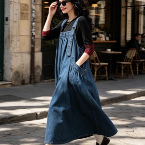 May include: A long blue denim overall dress with a gathered waist and side pockets, worn over a black long-sleeved shirt with red striped cuffs. The outfit is paired with white socks and dark brown leather loafers, creating a layered look.