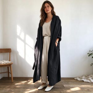 Puede incluir: Un abrigo largo tipo duster de color gris carbón oscuro, abierto por delante, con mangas largas y bolsillos laterales, sobre una camiseta sin mangas beige y pantalones beige a juego con cintura ceñida, junto a zapatos planos blancos.