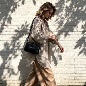 May include: A beige long-sleeved linen tunic with a V-neckline and curved hem, paired with loose-fitting tan trousers and a black leather shoulder bag with a flap closure and a thin strap, worn by a person with shoulder-length wavy blonde hair.