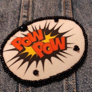 Può includere: Una toppa in tessuto di forma ovale con la scritta "POW POW" in lettere arancioni su un motivo a esplosione in stile fumetto giallo e nero. La toppa è bordata da una fila di piccole perline nere e decorata con perline nere sparse sulla superficie.