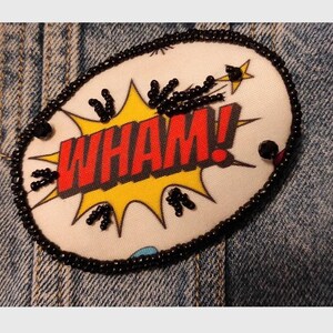 May include: An oval-shaped patch featuring a comic book style graphic with the word WHAM! in bold red letters against a yellow starburst background. The patch is outlined with black beads and includes additional black bead accents throughout.