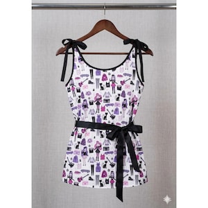 May include: A sleeveless white top featuring a repeating pattern of purple and black illustrations including figures, cats, guitars, and skateboards. The garment has black shoulder ties and a black waist sash tied in a bow at the front.