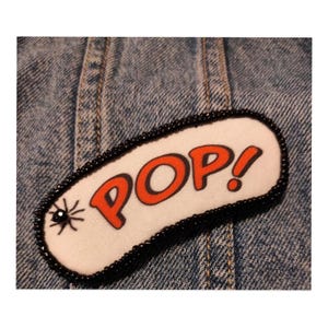 Può includere: Una toppa in tessuto di forma ovale con sfondo bianco e la parola POP! stampata in lettere arancioni vivaci con contorni neri. Il bordo è decorato con piccole perline nere e un piccolo disegno di un ragno nero si trova sul lato sinistro.