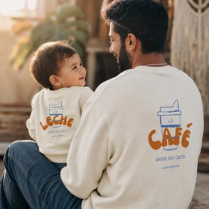 Könnte beinhalten: Zwei cremefarbene Sweatshirts mit Rundhalsausschnitt und blauen Strichzeichnungen von lächelnden Bechern. Das größere Sweatshirt zeigt den orangefarbenen Schriftzug CAFÉ, das kleinere LECHE, beide mit dem Text ANTES DEL CAOS CADA MAÑANA.