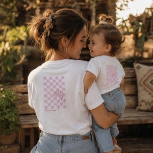 Könnte beinhalten: Zwei weiße T-Shirts mit einem rosafarbenen, karierten Rechteck-Motiv auf der Rückseite. Das Design zeigt eine Strichzeichnung einer Hand, die ein Croissant hält, sowie den Text "Mademoiselle croissant" in rosa Schreibschrift darunter.
