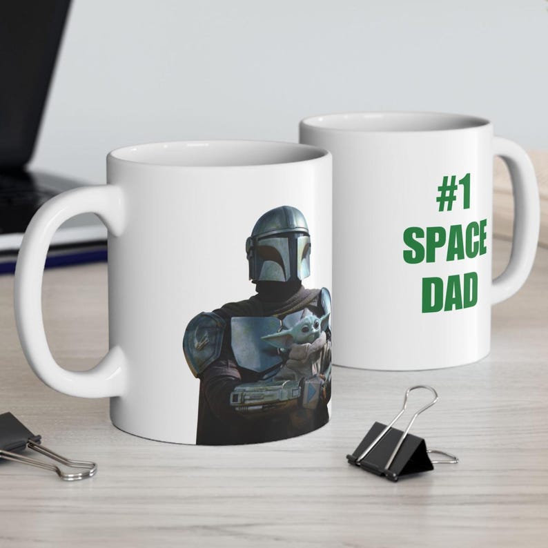 Puede incluir: Dos tazas de cer&aacute;mica blanca con un gr&aacute;fico de un personaje con armadura plateada que sostiene a una peque&ntilde;a criatura verde. Una taza muestra el texto #1 SPACE DAD en letras verdes. Las tazas tienen un asa est&aacute;ndar y una forma cil&iacute;ndrica.