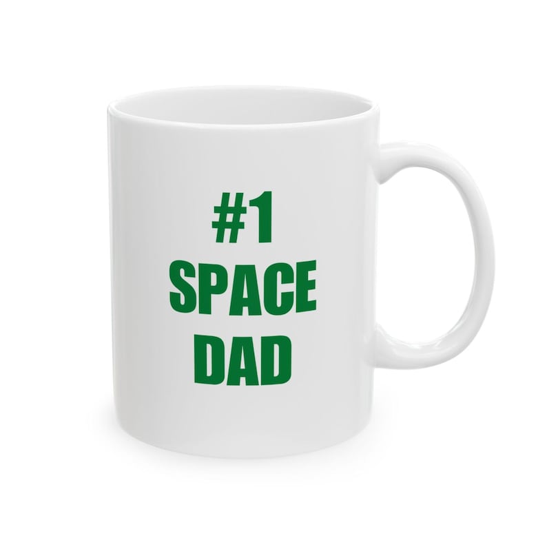 Puede incluir: Una taza de cer&aacute;mica blanca con el texto verde #1 SPACE DAD impreso en la parte frontal. La taza tiene un acabado liso y brillante, un cuerpo cil&iacute;ndrico y un asa curva en el lado derecho. El dise&ntilde;o est&aacute; centrado y utiliza una fuente negrita.