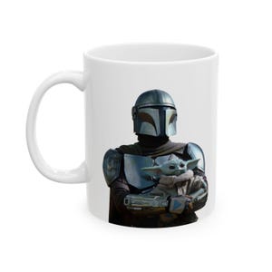 Puede incluir: Una taza de cer&aacute;mica blanca con un gr&aacute;fico impreso de un personaje con armadura plateada y casco, que sostiene a una peque&ntilde;a criatura verde de orejas grandes. El personaje lleva una armadura met&aacute;lica en el pecho y ropa oscura, la criatura viste una t&uacute;nica beige.