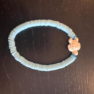 May include: A stretch bracelet composed of small, flat, circular light blue clay beads arranged in a continuous loop, featuring a single, textured, off-white turtle-shaped charm integrated into the design as a focal point of the accessory.