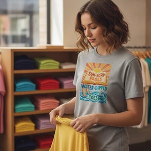 Peut inclure: Un t-shirt gris chiné à manches courtes avec une impression graphique représentant un livre ouvert sur fond de soleil et des vagues bleues en bas. Le texte sur le t-shirt indique SUN-KISSED PAGES et WATER-DIPPED TOES dans une police rétro colorée.