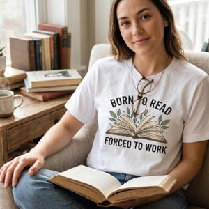 Peut inclure: Un t-shirt blanc à manches courtes avec un motif de livre ouvert entouré de feuilles vertes et le texte noir "BORN TO READ FORCED TO WORK" imprimé sur le devant. Le t-shirt est porté avec un jean bleu et des lunettes suspendues au col.