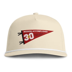 May include: A cream-colored baseball cap with a white cord across the brim. The front features a red pennant graphic with the text MISSION ACCOMPLISHED, the number 30, and the words MAJOR LEAGUE BALLPARK printed in white and black lettering.