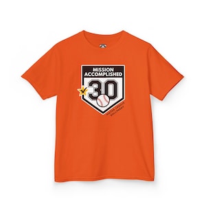 May include: A bright orange short-sleeve t-shirt featuring a graphic of a home plate shape with the text MISSION ACCOMPLISHED, the number 30, a baseball, a yellow star with a checkmark, and the text MAJOR LEAGUE BALLPARKS printed in black and white.