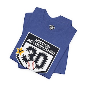 May include: A heather blue t-shirt featuring a graphic with the text "MISSION ACCOMPLISHED" above the number "30" in black font. A white baseball with red stitching sits at the base of the number, and a yellow star with a checkmark is included.