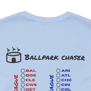 May include: A light blue t-shirt featuring a dark gray graphic of a stadium icon and the text "BALLPARK CHASER" in a bold font. Below the text are two columns of circular checkboxes next to red and blue baseball team abbreviations.