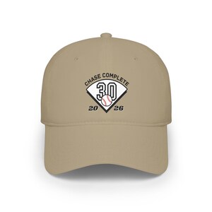 May include: A tan baseball cap featuring a graphic of a home plate with the number 30, a baseball, and the text CHASE COMPLETE 20 26. The design is centered on the front panel of the cap, which has a curved brim and a structured crown.