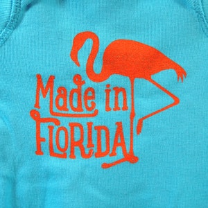 Puede incluir: Un body turquesa con un flamenco naranja y el texto "Made in Florida". El texto y el flamenco est&aacute;n impresos en negrita. El body est&aacute; hecho de una tela suave y acanalada.