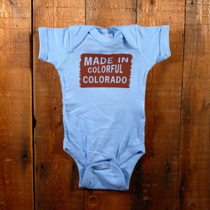 May include: A light blue baby onesie with short sleeves. The onesie has the text "MADE IN COLORFUL COLORADO" in a brown rectangular shape on the front. The onesie is made of soft material and has snap closures at the bottom.