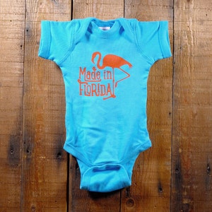 Puede incluir: Un body de beb&eacute; turquesa con un gr&aacute;fico de flamenco naranja brillante y el texto "Made in FLORIDA". El body est&aacute; hecho de material suave y est&aacute; colocado sobre una superficie de madera.