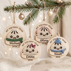 Puede incluir: Cuatro adornos navideños redondos de madera colgados de una rama de pino. Cada uno presenta un personaje de bola de masa brillante en una vaporera con el texto 'MY SQUISHY YEAR', un nombre y '2026'. Las bolas de masa llevan bufandas, gorros o lazos.