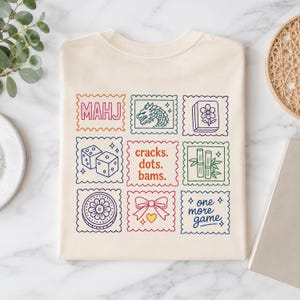 Pu&ograve; includere: Una t-shirt color crema con una griglia di nove grafiche quadrate dai bordi smerlati. I disegni includono il testo "MAHJ", un drago, una tessera fiore, due dadi, il testo "cracks. dots. bams.", bamb&ugrave;, un motivo circolare, un fiocco e "one more game."