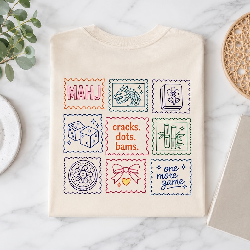 Pu&ograve; includere: Una t-shirt color crema con una griglia di nove grafiche quadrate dai bordi smerlati. I disegni includono il testo "MAHJ", un drago, una tessera fiore, due dadi, il testo "cracks. dots. bams.", bamb&ugrave;, un motivo circolare, un fiocco e "one more game."
