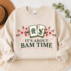 May include: A beige crewneck sweatshirt featuring a graphic of two mahjong tiles surrounded by pink cherry blossoms. The text reads IT'S ABOUT BAM TIME in dark green capital letters, with a small pink heart icon centered below the text.