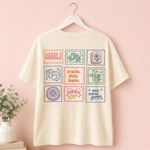 Pu&ograve; includere: Una t-shirt a maniche corte color crema con una griglia di nove grafiche quadrate colorate sul retro. I disegni includono icone a tema Mahjong come un drago, bamb&ugrave;, dadi, una tessera fiore, un fiocco e il testo MAHJ, cracks. dots. bams. e one more game.