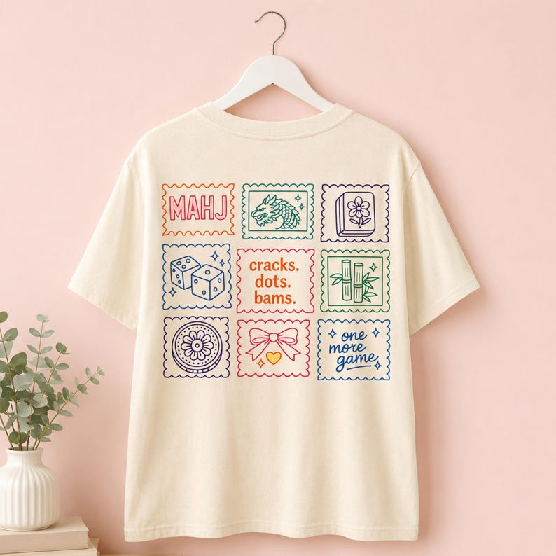 Pu&ograve; includere: Una t-shirt a maniche corte color crema con una griglia di nove grafiche quadrate colorate sul retro. I disegni includono icone a tema Mahjong come un drago, bamb&ugrave;, dadi, una tessera fiore, un fiocco e il testo MAHJ, cracks. dots. bams. e one more game.