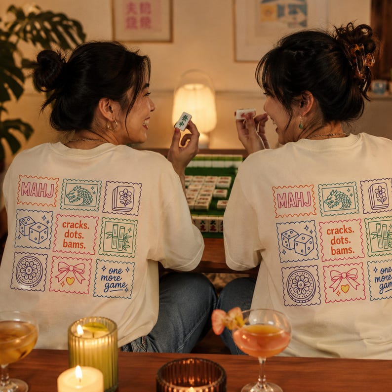 Pu&ograve; includere: Una t-shirt color crema con una griglia di nove quadrati colorati in stile ricamo sul retro. I quadrati contengono illustrazioni a linee di tessere del mahjong, dadi, bamb&ugrave;, un fiore, un fiocco e il testo MAHJ e one more game.