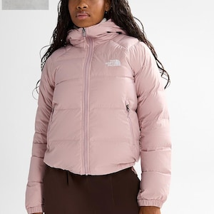 May include: A light pink puffer jacket with a hood, a full-length front zip, and two zipped side pockets. The North Face logo is embroidered in white on the upper left chest. The jacket features horizontal quilted stitching and elasticated cuffs.