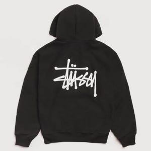 May include: A black long-sleeve hooded sweatshirt featuring a white Stüssy logo printed on the back. The garment includes a hood, long sleeves with ribbed cuffs, and a ribbed hem, showcasing the brand name in a stylised, handwritten font.