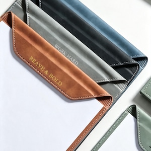 May include: A collection of document folders in tan, gray, and blue, each featuring white stitching along the edges. The tan folder is embossed with the text BRAVE & BOLD, while the gray folder displays the text WORK HARD in a clean, sans-serif font.