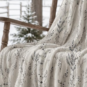 May include: A cream-coloured throw blanket featuring a dark grey botanical print of wildflowers and grasses draped over a wooden rocking chair. The fabric displays a repeating pattern of delicate stems, leaves, and seed heads across the surface.