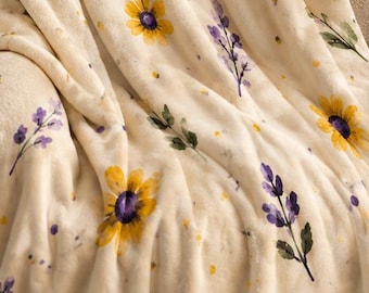 May include: A cream-colored plush throw blanket featuring a repeating pattern of yellow sunflowers and purple lavender sprigs with green stems. The soft fabric is draped over a chair, displaying the floral print across the entire surface area.