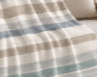 May include: A soft throw blanket featuring horizontal stripes in shades of muted blue, light gray, and beige against a white background. The blanket is draped over a neutral-colored armchair, showcasing a watercolor-like texture across the fabric.