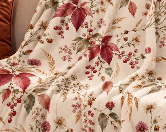 May include: A cream-colored throw blanket featuring a repeating botanical print of deep red autumn leaves, small red berries, and dried golden wheat stalks. The fabric has a soft texture and is draped over the back and seat of a neutral sofa.