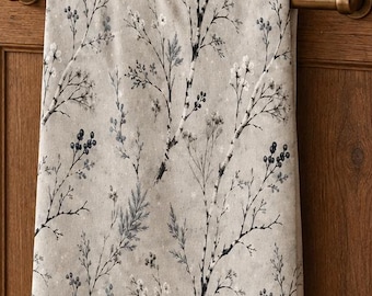 May include: A light gray kitchen towel featuring a dark blue and white botanical print of winter branches, berries, and small flowers. The rectangular towel hangs vertically over a horizontal brass handle attached to a wooden kitchen cabinet door.