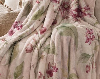 May include: A soft, plush throw blanket featuring a light pink base color with a watercolor-style floral pattern in shades of deep magenta, burgundy, and muted green leaves. The blanket is draped over a light-colored upholstered armchair.
