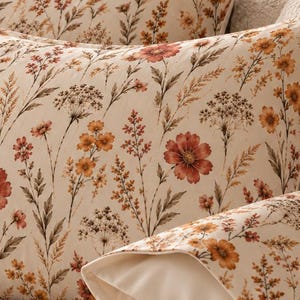 May include: Two rectangular bed pillows featuring a cream-coloured fabric printed with a botanical pattern of wildflowers in shades of rust orange, muted yellow, and brown, accented by thin green stems and delicate foliage on a textured bed.