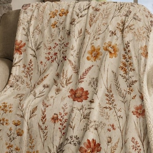 May include: A cream-colored throw blanket featuring a botanical print of wildflowers in shades of orange, rust, and brown. The blanket has a soft, textured fleece-like surface on one side and a thick, white sherpa lining on the reverse side.