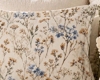 May include: A rectangular pillow sham and a matching quilted bedspread featuring a botanical print of delicate blue and golden-brown wildflowers on an off-white background. The bedding set is displayed on a bed with textured cream-colored layers.